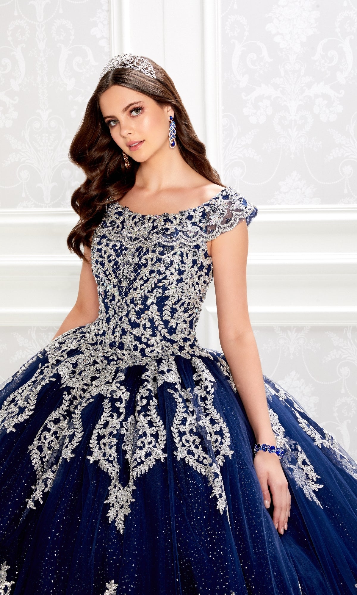 A young woman in the PR22035 Quinceanera Dress by Princesa, featuring silver embroidery, glitter tulle, and a corset bodice, stands against an ornate white background wearing a jeweled tiara.